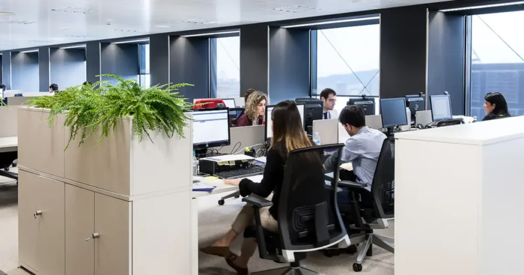 Oficina diáfana con falso techo y rejillas lineales de climatización sobre los puestos de trabajo, donde varios empleados trabajan frente a sus ordenadores.