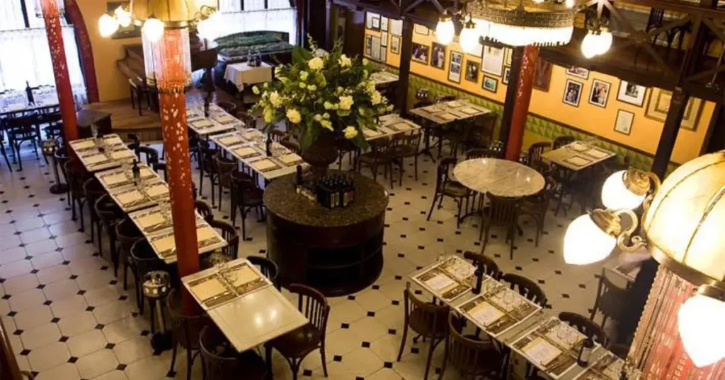 Comedor de un restaurante clásico visto desde arriba, con mesas montadas, suelo de baldosas blancas y negras, columnas rojas y lámparas de techo encendidas.